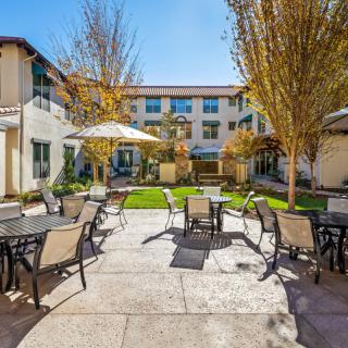 A sunny courtyard with outdoor seating and tables, surrounded by a building and trees.