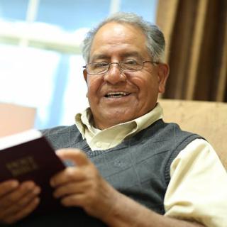 An elderly man smiling while reading a book indoors.