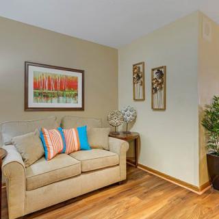 A cozy living room corner featuring a beige sofa with colorful pillows, artwork on the walls, and green plants adding a touch of nature.