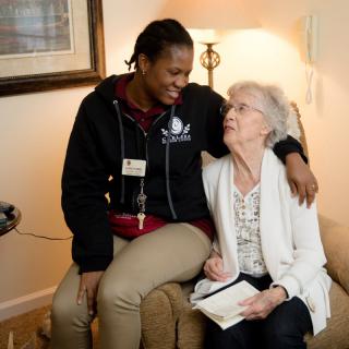 An elderly woman and a caregiver share a warm moment together, smiling in a comfortable setting.