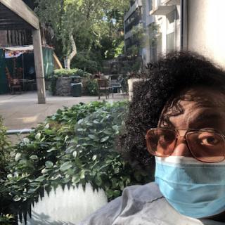 A person wearing a mask takes a selfie in front of a window, showcasing a vibrant outdoor scene with greenery.