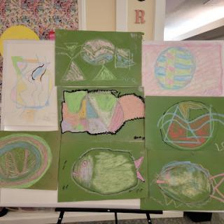 A collection of colorful children's artwork displayed on a board, featuring various abstract designs against green backgrounds.