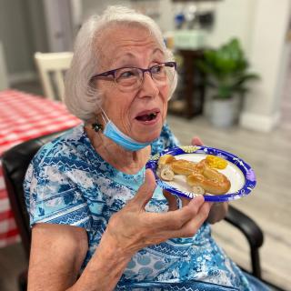 An elderly woman enjoys a meal indoors while wearing a mask under her chin.