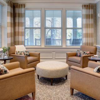 A cozy living room with comfortable chairs arranged around a central ottoman, large windows, and stylish curtains.