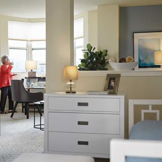 A cozy living space featuring a dresser with a lamp, artwork on the wall, and a dining area in the background.