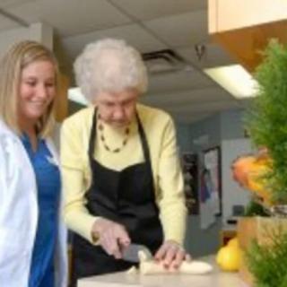 An elderly woman slices food alongside a young woman in a kitchen setting.