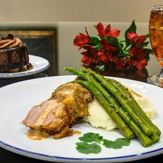 A delicious dinner plate featuring pork with gravy, asparagus, and mashed potatoes, accompanied by a dessert cake and a drink.