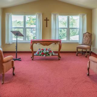 A small chapel with a cross, chairs, and a table in front of large windows.