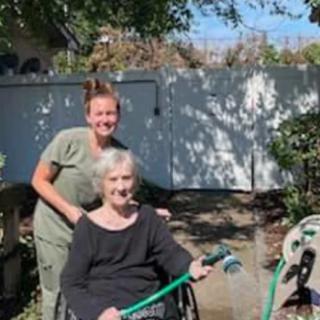 An elderly person in a wheelchair holding a garden hose, accompanied by a younger caregiver, enjoying a sunny day in the garden.