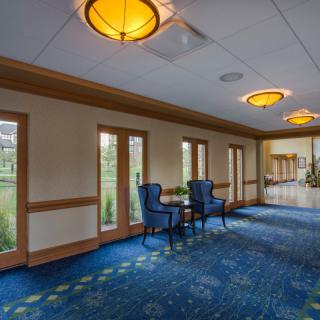 A spacious hallway with blue carpet and elegant lighting, featuring a seating area by large windows.