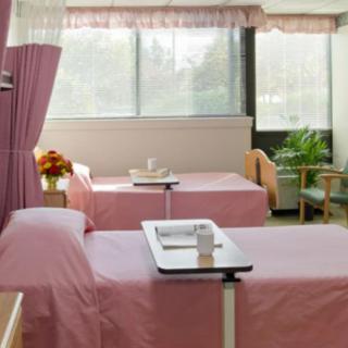 A hospital room with two patient beds covered in pink linens, separated by a curtain, and brightened by natural light from large windows.