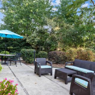 A cozy patio with outdoor seating, surrounded by lush greenery and shaded by an umbrella.