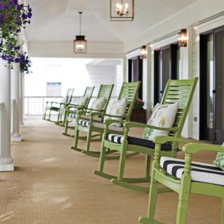 A row of green rocking chairs with striped pillows line a porch, illuminated by hanging lanterns.