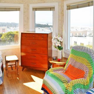 A cozy living room with an armchair covered in a colorful blanket, wooden furniture, and large windows offering a scenic view.
