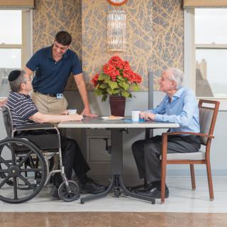 A caregiver interacting with two elderly individuals at a table in an indoor setting.