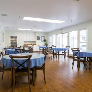 A brightly lit dining room with multiple tables covered in blue tablecloths and wooden chairs.
