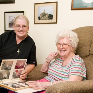 An elderly woman and her caregiver are happily looking through a photo album in the living room.