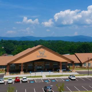 A school building with a parking lot, surrounded by trees and mountains under a blue sky with clouds.