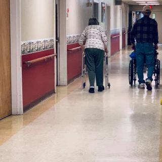 Elderly individuals walking down a nursing home hallway with assistance.