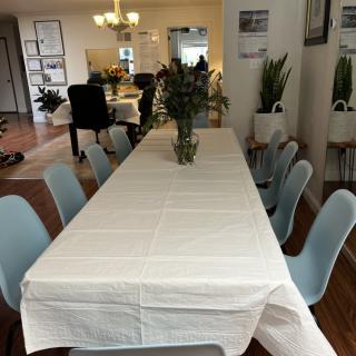 A dining room setup featuring a long table with a white tablecloth, surrounded by chairs and adorned with plants.