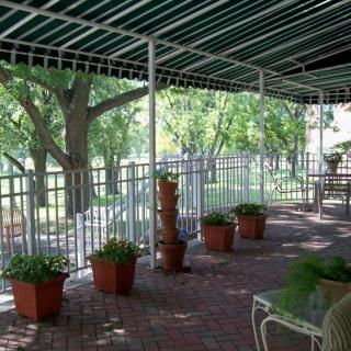 A shaded patio with potted plants and outdoor furniture, surrounded by trees.