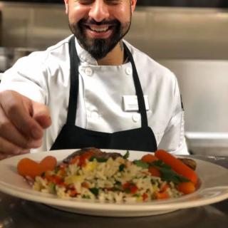 A smiling chef presenting a beautifully garnished plate of food in a kitchen.