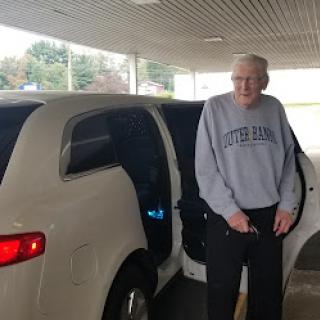 An elderly man standing beside a car, ready to embark on his journey.