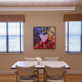 A dining room featuring a white table and chairs, flanked by two windows with an abstract painting on the wall.