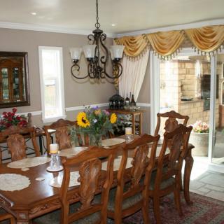 A spacious dining room featuring a large wooden table set with chairs, an ornate chandelier, and elegant curtains framing sliding doors. A vase of sunflowers adds a touch of color to the classic setting.
