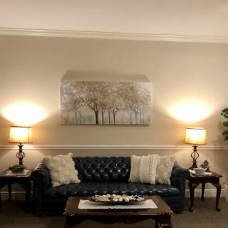 A cozy living room with a blue sofa, illuminated by two lamps, adorned with pillows and artwork of trees on the wall.