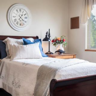 A cozy bedroom with a neatly made bed, adorned with decorative pillows and a nearby nightstand featuring a lamp and flowers.
