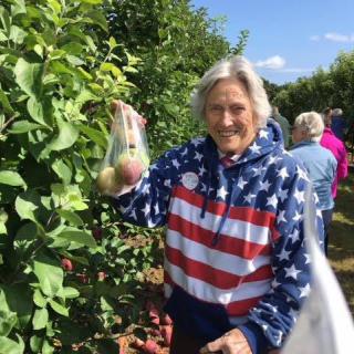 A woman wearing a flag-patterned jacket is apple picking in a garden.