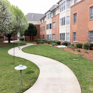 A curved pathway through a landscaped area with lawn and trees, adjacent to a brick building.