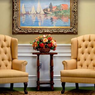 A cozy setting with two upholstered chairs and a floral arrangement, set against a backdrop of a framed painting depicting sailboats on water.