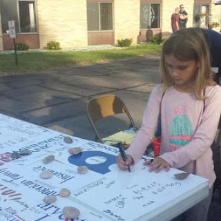 A young girl is engaged in signing or drawing on a large table at an outdoor event.