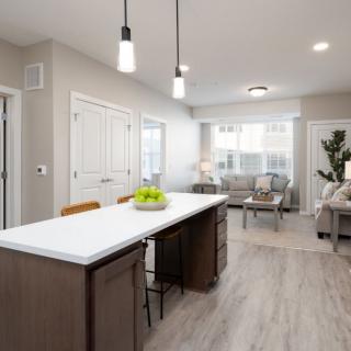A modern kitchen with an island counter opens into a cozy living area, featuring minimalist interior design and ample natural light.