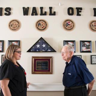 Two individuals stand in front of a wall dedicated to honoring veterans, adorned with military insignia and an American flag.