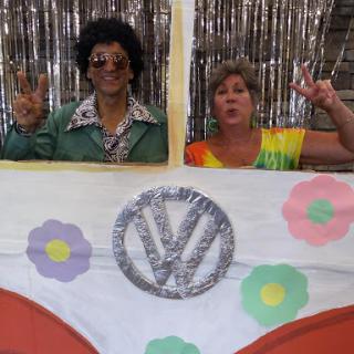 Two individuals dressed in vibrant 70s-style costumes pose playfully behind a decorated Volkswagen-themed backdrop.