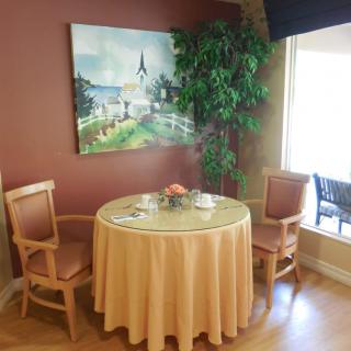 A cozy dining nook with a round table set for two, adorned with a peach tablecloth and complemented by elegant chairs. An indoor plant and a landscape painting enhance the atmosphere, while natural light streams in through a nearby window.