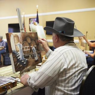 An artist in a hat painting on a canvas during an art class.