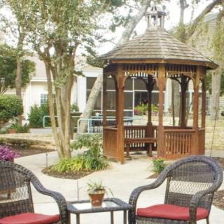 A tranquil garden setting featuring a wooden gazebo with surrounding chairs and lush greenery.