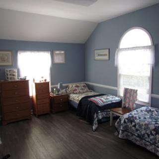 A cozy bedroom with blue walls features two twin beds, a dresser, and ample natural light from large windows.