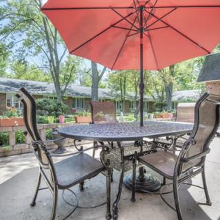 A cozy patio setup with a table, chairs, and a red umbrella in a peaceful garden setting.