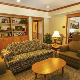 A cozy living room with a floral sofa, two armchairs, a wooden coffee table, bookshelves, and a warm lamp.