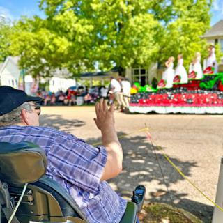 A person in a wheelchair enjoys the festivities of a vibrant parade, capturing the moment with their phone.