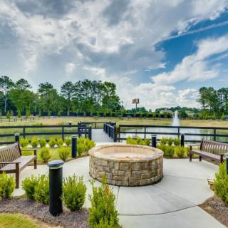 A scenic outdoor area featuring a stone fire pit surrounded by benches, overlooking a pond with a fountain under a cloudy sky.
