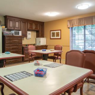 A cozy room with multiple tables and chairs, featuring a kitchenette area with cabinets and a window.