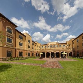 A view of a large building with a courtyard and garden under a partly cloudy sky.