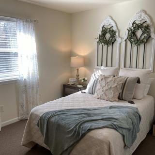 A cozy and well-decorated bedroom with a bed adorned with pillows and a decorative headboard, gently lit by natural light from the window and a lamp on the nightstand.