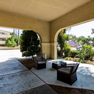 A covered outdoor patio area with comfortable seating and a table, surrounded by greenery on a sunny day.